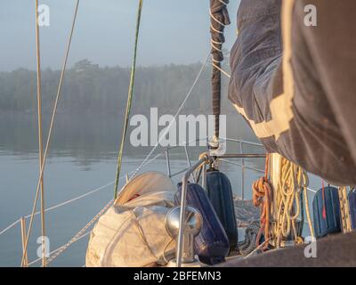 Ich freue mich auf den Bug eines Segelbootes im stillen Wasser mit den Segeln nach unten und Ausrüstung auf Deck mit Blick auf eine dunstbewaldete Küste, die von Nebel verdeckt ist. Stockfoto