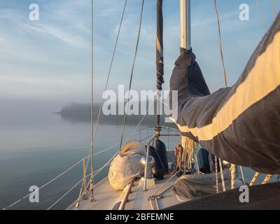 Bug eines Segelbootes an einem ruhigen, nebligen Tag mit Segeln bedeckt und Ausrüstung auf Deck mit Blick auf einen dunstigen bewaldeten Punkt, wo der Horizont verschwindet im Nebel. Stockfoto