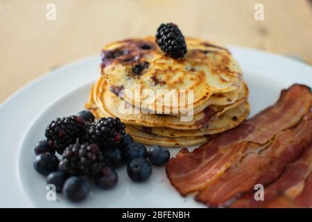 Frühstück mit Heidelbeer-Pfannkuchen mit Speck, Obst und Ahornsirup auf einem weißen Teller serviert. Stockfoto