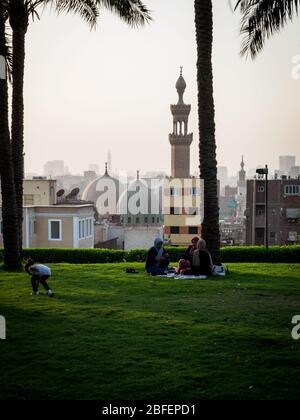 Al Azhar Park, Kairo, Ägypten, Oktober 2019, drei Mädchen sitzen auf dem Gras und ein Kind spielen mit der Skyline von kairo im Hintergrund Stockfoto