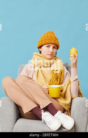 Porträt der jungen Frau in Wollmütze und Schal sitzen im Sessel und trinken heißen Tee auf blauem Hintergrund Stockfoto