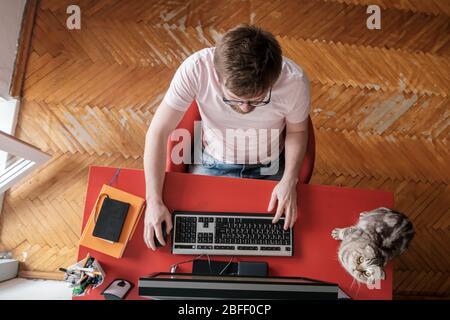 Mann arbeitet ferngesteuert zu Hause auf einem Computer, neben sitzt seine liebenswerte Katze. Konzept der Selbstisolierung während einer Pandemie. Lifestyle. Stockfoto