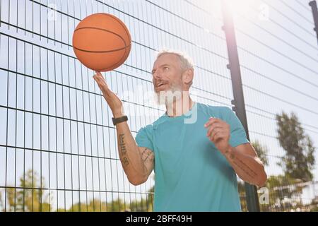 Basketball im Freien spielen. Glücklich älteren Mann in Sportkleidung Spinnen einen Basketballball auf seinem Finger, während auf Outdoor-Basketballplatz stehen Stockfoto