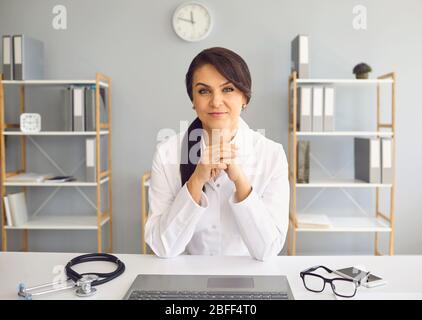 Praktizierende Ärztin, die die Kamera anschaut, während sie an einem Tisch in einem Klinikbüro sitzt. Stockfoto