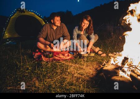 Lächelndes fröhliches Paar, das vor einem kochenden Abendessen sitzt. Camping-Konzept Stockfoto