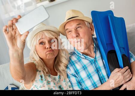 Senior paar sitzt in Reisebüro sitzen mit Hüten Mann hält Flossen, während Frau Selfie-Bilder auf Smartphone Grimace lustig Stockfoto