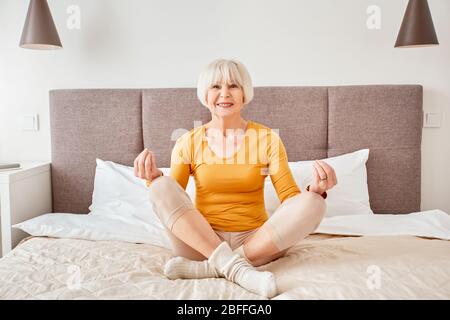 Reife ältere Frau sitzt in Meditationsose auf dem Bett Stockfoto