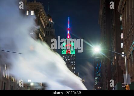 Empire State Building leuchtet in Weihnachtsfarbe hinter dem treibenden Dampf zwischen den Fifth Avenue Gebäuden in der Nacht in New York NY USA am 2018. Dezember Stockfoto