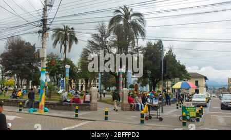 Cayambe, Pichincha / Ecuador - Februar 24 2020: Menschen sitzen in der Ecke des zentralen Parks der Stadt mit der Matriz-Kirche im Hintergrund Stockfoto