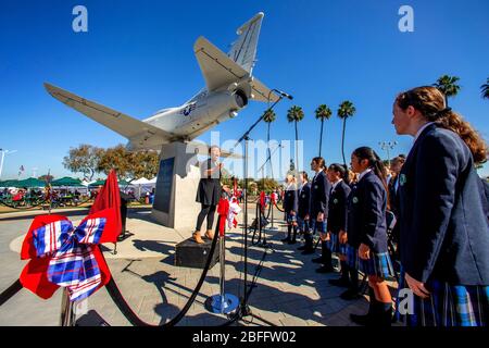 Ein katholischer Mädchenchor wird von ihrem afroamerikanischen Führer geleitet, während sie patriotische Lieder bei Veterans Day Observances in Costa Mesa, CA singen. Die A4M Skyhawk ist ein patriotisches Denkmal. Stockfoto