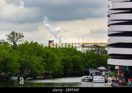 Blick auf weißen Rauch steigt aus einem Kamin einer Fabrik mit Blick auf einen Kanal in Amsterdam, Niederlande Stockfoto