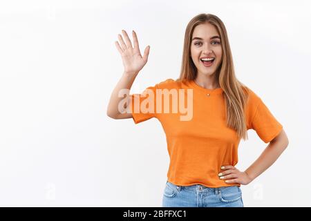 Hallo Nachbar. Lächelndes blondes Mädchen, das zu Hause in der Nähe des Fensters steht und ihren Freund über die Straße winkt, um Hallo zu sagen, Gruß Stockfoto