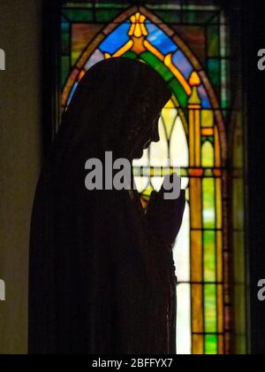 Eine Statue der Jungfrau Maria beim Gebet ist gegen ein Buntglasfenster in einer katholischen Kirche in Twin Mountain, New Hampshire silhouetted. Stockfoto