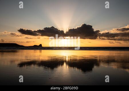 Sonnenuntergang am Strand von Rhossili Bay mit Wolkenreflexionen und Sonnenstrahlen hinter den Wolken, die auf Worm's Head on the Gower, South Wales, blicken. GROSSBRITANNIEN Stockfoto