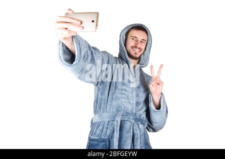 Porträt des jungen kaukasischen bärtigen Mannes in blauem Bademantel zeigt Sieg Geste und macht Selfie isoliert auf weißem Hintergrund. Happy Emotion Konzept Stockfoto