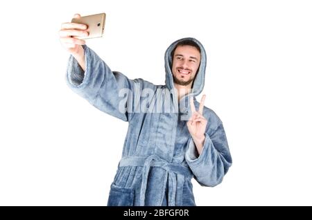 Porträt des jungen kaukasischen bärtigen Mannes in blauem Bademantel zeigt Sieg Geste und macht Selfie isoliert auf weißem Hintergrund. Happy Emotion Konzept Stockfoto