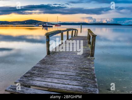 Ein alter Holzsteg und sichere, festgefahrene Boote tragen zum malerischen Sonnenuntergang auf der spiegelartigen Oberfläche der St Helen''s Bay in Tasmanien bei. Stockfoto