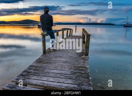 Touristen sitzen auf einem alten Holzsteg und sehen den perfekten Sonnenuntergang am Hafen, mit Booten, die sicher in St Helen''s Bay in Tasmanien verankert sind. Stockfoto