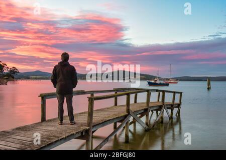 Touristen stehen auf einem alten hölzernen Steg und sehen den perfekten Sonnenuntergang am Hafen, mit Booten sicher in St Helen''s Bay in Tasmanien verankert. Stockfoto