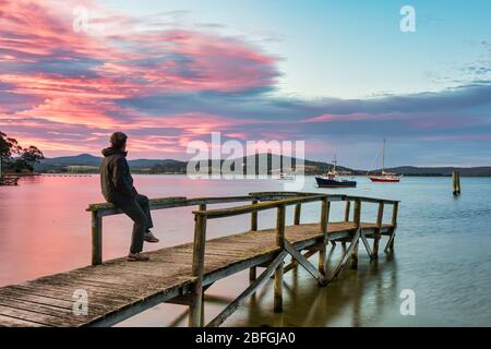 Touristen sitzen auf einem alten Holzsteg und sehen den perfekten Sonnenuntergang am Hafen, mit Booten, die sicher in St Helen''s Bay in Tasmanien verankert sind. Stockfoto