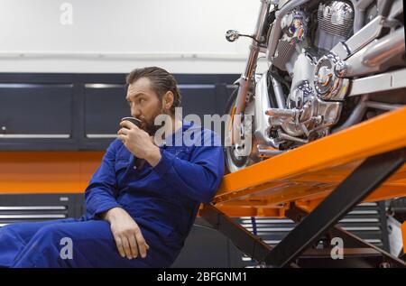 Ein Mann in der Werkstatt sitzt neben einem Motorrad und trinkt Kaffee. Nach Abschluss der Reparatur mit Motorrad-Wartung ausruhen. Stockfoto