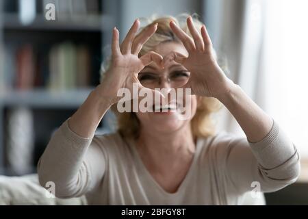 Ältere Frau auf der Couch sitzend, die das Herzsymbol mit den Fingern zeigt Stockfoto