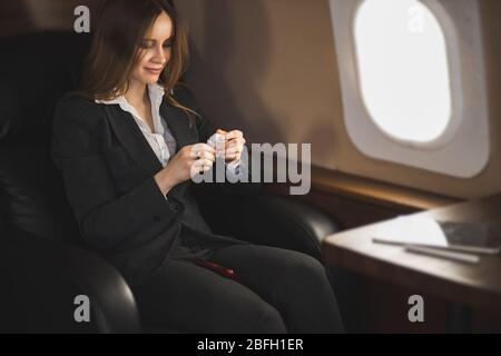 Attraktive kaukasische junge Frau Unternehmerin sitzt in bequemen Sitz in Privatflugzeug, lächelnd mit modernen kabellosen Kopfhörer, gehen zu li Stockfoto