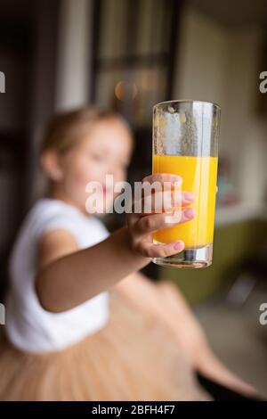 Nettes kleines kaukasisches Mädchen mit blonden Haaren, das auf dem Küchentisch sitzt und frischen Mangosaft trinkt. Gesundes Lifestyle-Konzept. Kind 6 Jahre alt bei Stockfoto