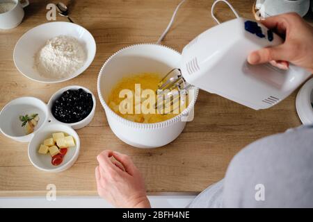 Junge Frau, die in der Küche einen Edelsteinkuchen backen kann, steht an der Theke und bringt mit einem Handmixer frische Zutaten in eine weiße Rührschüssel. Stockfoto