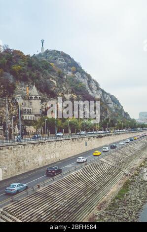 Budapest, Ungarn - 6. November 2019: Gellert Hill Cave Church, Sziklatemplom. Freiheitsstatue auf dem Hügel. Autos auf der Straße vor dem Hügel an der Donau. Vertikales Foto. Stockfoto