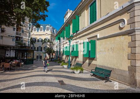 Einheimische Frau, die auf einer engen Kopfsteinpflasterstraße im historischen Viertel von Macau, China, spazieren geht. Stockfoto