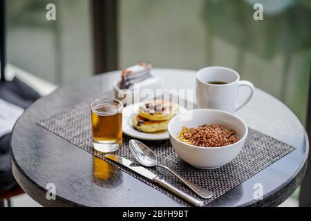 Ein Frühstücksset: Eine Schüssel Müsli, Pfannkuchen, eine Tasse Kaffee und Apfelsaft. Stockfoto