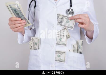 Cropped Nahaufnahme Foto von Frau Doktor versteckt Papiergeld Banknoten unter ihrem weißen Mantel und in Taschen isoliert über grauem Hintergrund Stockfoto