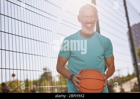 Glücklich ältere Basketballspieler in Sportkleidung, die Basketball hält und lächelt an der Kamera, während sie auf Outdoor-Basketballplatz steht Stockfoto