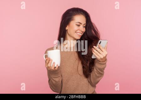 Porträt von stilvollen attraktive junge Frau mit langen braunen Haaren hält Tasse Kaffee und Nachricht auf dem Smartphone beobachten, mit Pause und entspannend. ich Stockfoto