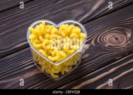 Rohe Pasta auf dunklen Holzplanken Hintergrund. Lakonisches Design. Symbol der Liebe. Minimalistischer Stil. Gelb rohe Makkaroni in Schale Herz auf einem braunen geformt Stockfoto