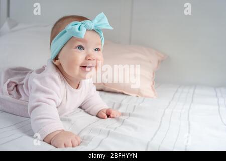 Neugeborenes liegt auf dem Bauch und lächelt. Ein Kind mit blauen Augen und blauem Stirnband. Niedliches Baby trägt auf dem Bauch im Kinderzimmer liegen. Wunderschöne s Stockfoto