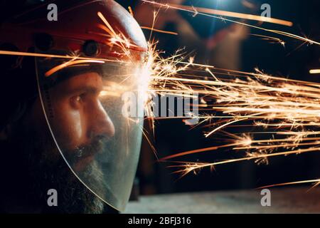 Mann Arbeiter in transparenter Schutzmaske arbeitet auf Metall mit Kreissäge Schleifer. Funken und Gesicht. Stockfoto