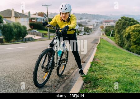 18/04/2020-Asiatische Frau macht bergauf mit Mountainbike. Eine Frau, die sehr müde war und auf der Straße in Oamaru, Neuseeland, anhält. Stockfoto