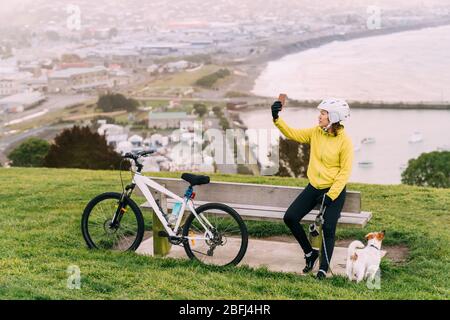 Asiatische Frau machen bergauf mit Mountainbike. Eine Frau, die ein Foto am Aussichtspunkt mit einem Hund in Oamaru, Neuseeland, macht. Stockfoto