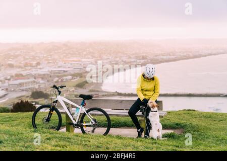 Asiatische Frau machen bergauf mit Mountainbike. Eine Frau, die mit einem Hund am Aussichtspunkt Oamaru, Neuseeland, spielt. Stockfoto