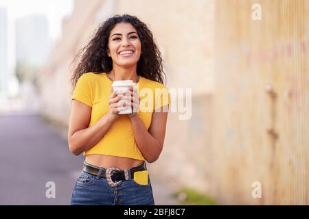 Arabisches Mädchen, das mit einem Kaffee zum Mitnehmen auf der anderen Straßenseite spazieren geht Stockfoto