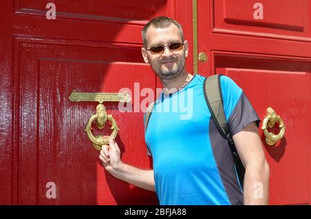 Ein Tourist mittleren Alters klopft an die rote Tür mit einem dekorativen Türrapper und goldenem Buchstabenschlitz Stockfoto