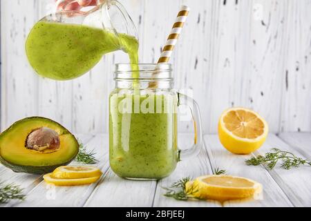 Ein gesunder grüner Smoothie wird aus einer Kanne in ein Glas gegossen. Grüner Smoothie mit Bio-Zutaten, Gemüse auf weißem Holzhintergrund. Stockfoto