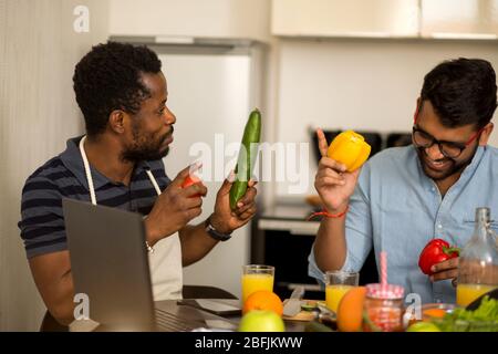 Schönen afrikanischen amerikanischen Jungen Mann und seiner indischen Freund am Tisch Gemüse Holding sitzen, diskutieren, Rezept, an jedem anderen suchen, mit Laptop Stockfoto