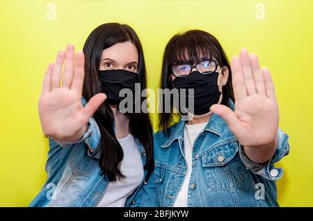 COVID-19. Mutter und Tochter in schützender medizinischer Gesichtsmaske zeigen Handzeichen-Stop auf gelbem Hintergrund. Anti-Coronavirus-Konzept. Stockfoto