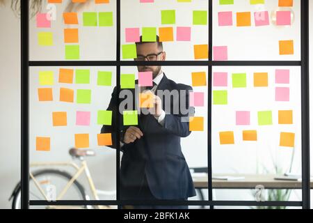 Konzentrierter Geschäftsmann schreibt auf Haftnotizen auf Glas Bürowand Stockfoto