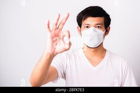 Nahaufnahme Asian Handsome Mann trägt chirurgischen hygienischen Schutztuch Gesichtsmaske gegen Coronavirus und zeigen OK Zeichen, Studio erschossen isoliert weißen Rücken Stockfoto