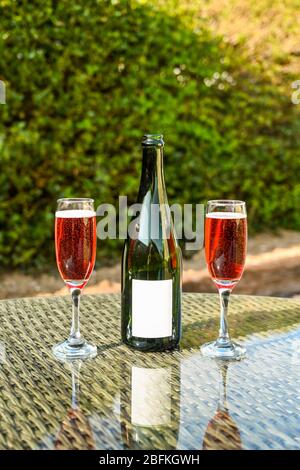 Zwei Flötengläser aus rosa Champagner und eine geöffnete Champagnerflasche mit einem leeren Etikett auf einer Glastisch-Platte in einem Garten. Platz für Kopie. Stockfoto