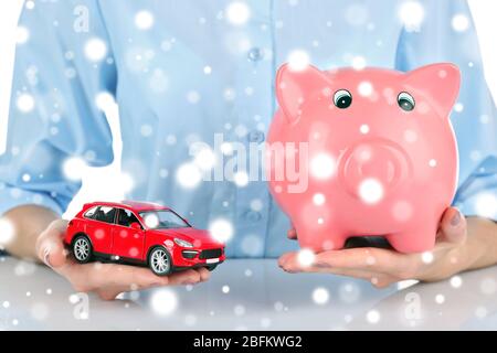 Weibliche Hände halten Sparschwein und Modell des Autos über Schnee-Effekt Stockfoto